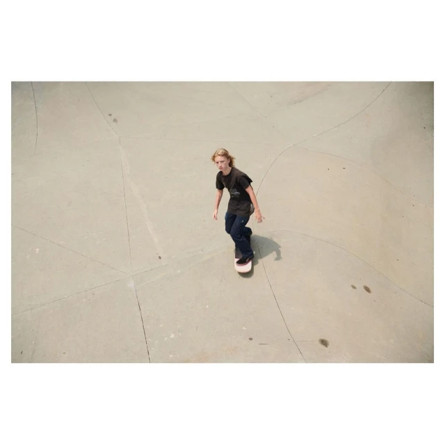 Young adult with long blond hair rides a skateboard in an outdoor concrete skatepark, shot from a high angle, capturing the sport's action.