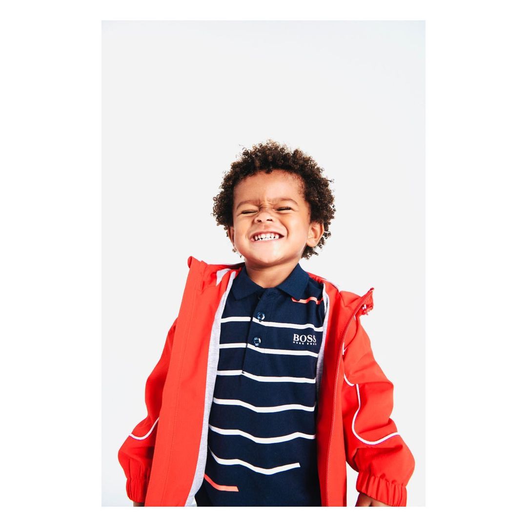 Portrait of a young African-descent boy with curly hair, wearing a navy and white striped polo shirt and a red jacket, smiling at camera.