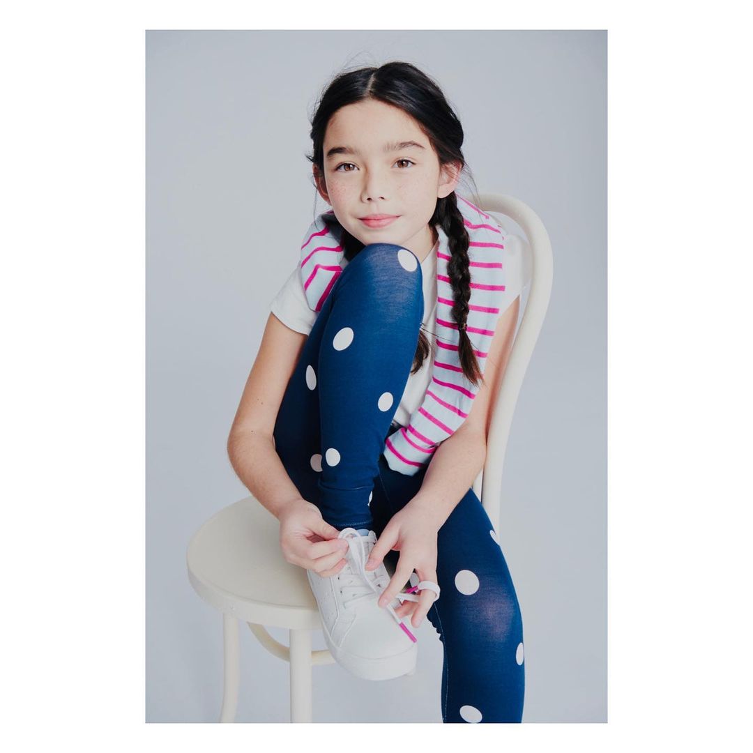 A girl with braided hair in blue polka dot leggings sitting on a chair and tying the laces of her white sneakers in a light background.