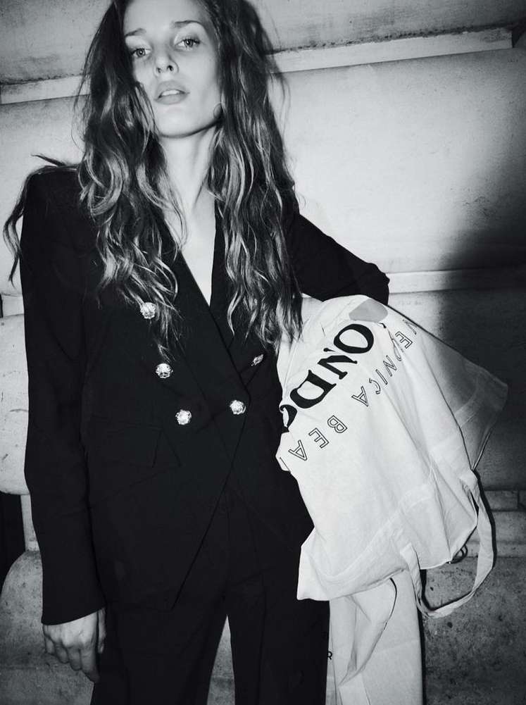 Black and white portrait of a woman in a dark double-breasted blazer holding a printed tote bag against stone architecture.