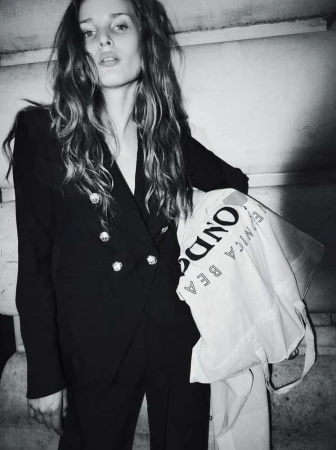 Black and white portrait of a woman in a dark double-breasted blazer holding a printed tote bag against stone architecture.
