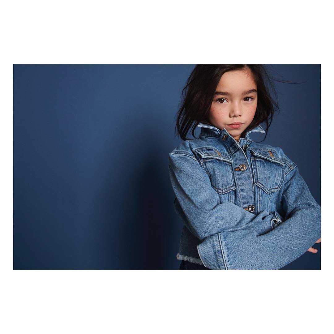 Portrait of a young girl with dark hair, wearing a light-blue denim jacket against a solid dark-blue background. Studio shot.