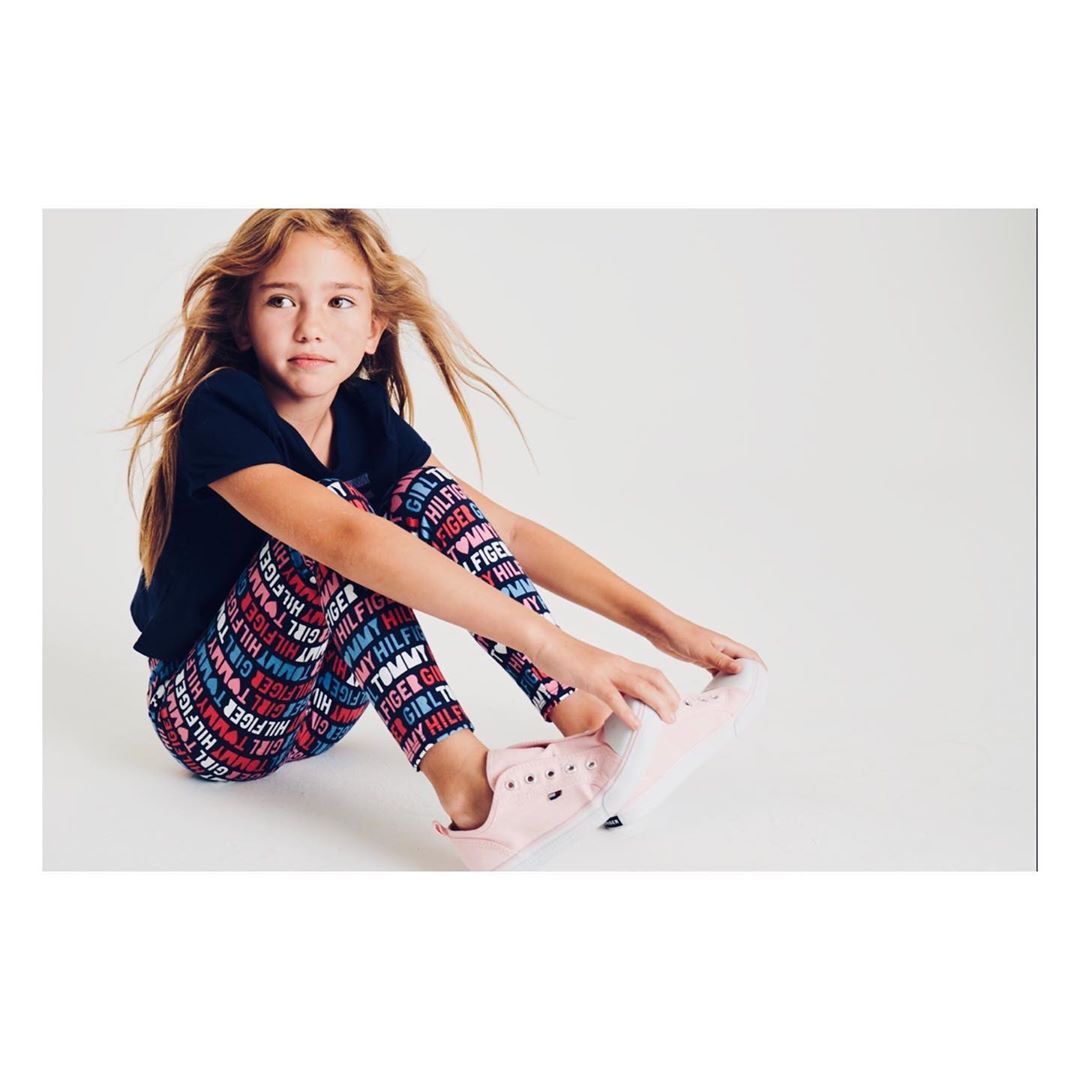 Young girl seated in photo studio wears Tommy Hilfiger logo leggings with pink sneakers and a navy t-shirt against a white backdrop.
