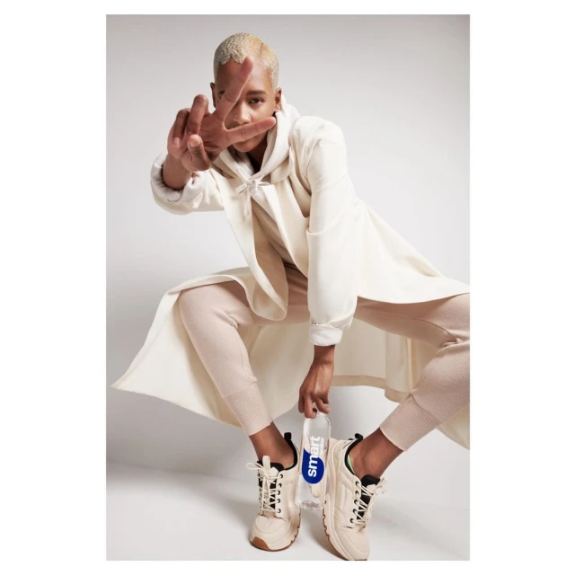 Stylish young adult in a crouched pose, wearing a cream-colored outfit and sneakers, holds a Smartwater bottle in a bright studio setting.