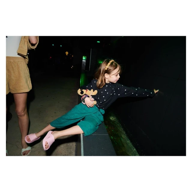 Child in star-patterned shirt and green culottes sitting with a stuffed moose in a dark indoor space, with a woman nearby.