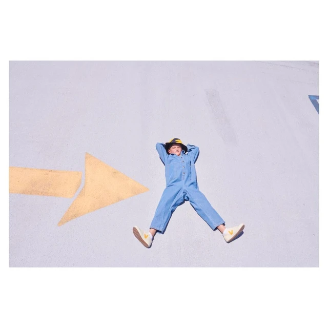 A child in a blue jumpsuit and hat lying on a gray surface with a yellow arrow, arms behind their head, legs spread in sunlit outdoor shot.
