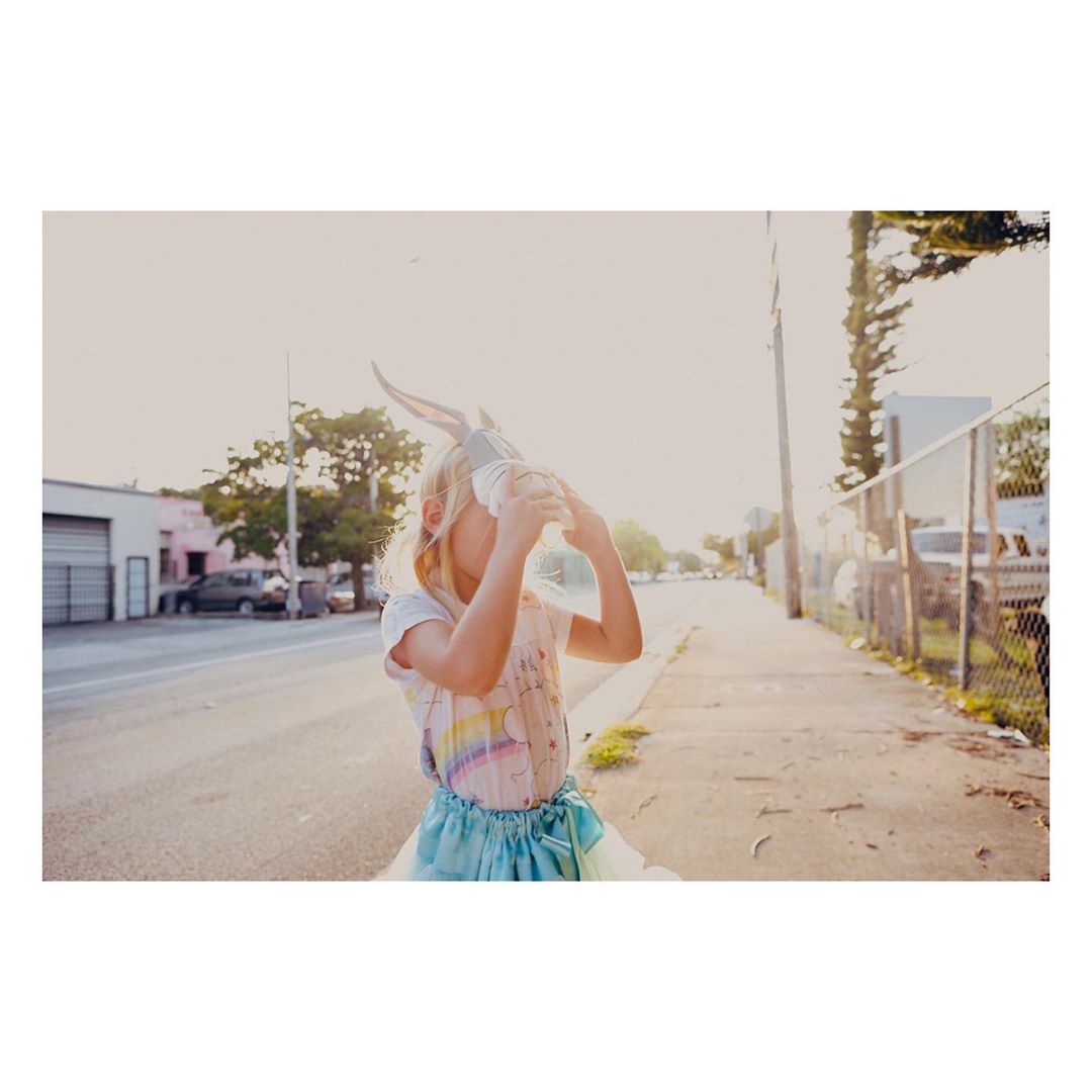 Young girl wearing bunny ears drinking from a clear plastic bottle outdoors on a sunny street with buildings and trees in the background.
