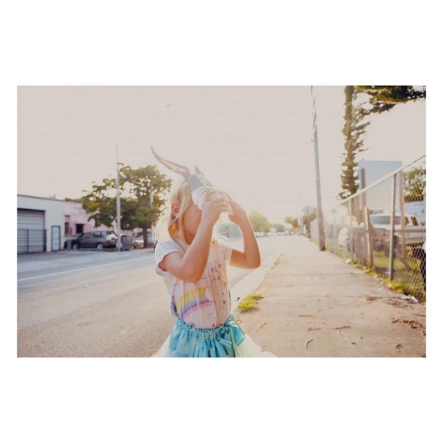 Young girl wearing bunny ears drinking from a clear plastic bottle outdoors on a sunny street with buildings and trees in the background.