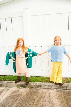 Two young girls hold hands wearing retro-style clothes in front of a white fence on a sunny day. One wears a pink dress and the other yellow pants.