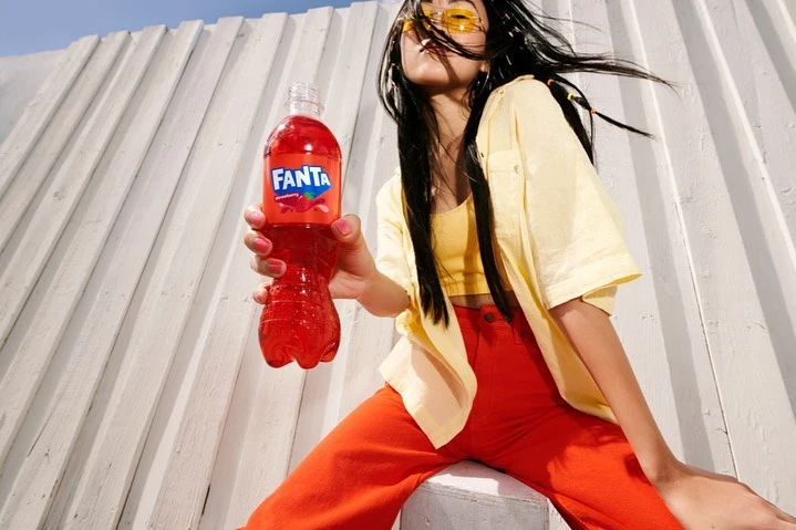 Young woman in trendy outfit sits near a textured wall, holding a bottle of Fanta soda. Bright sunlight and vibrant colors.