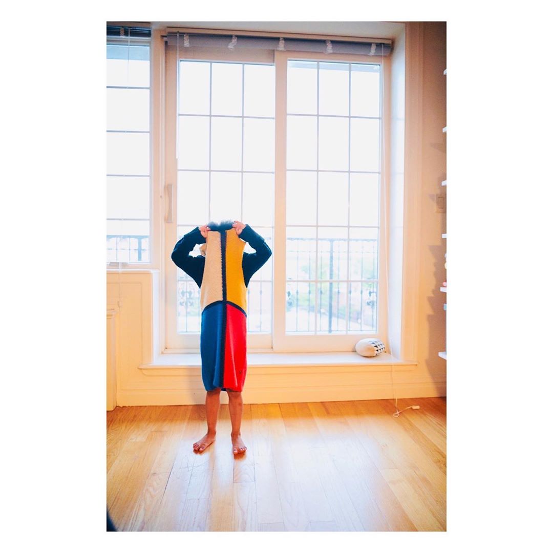 Child stands in a room with wooden floors wearing a colorful homemade Mondrian-inspired dress in front of a large paned window.