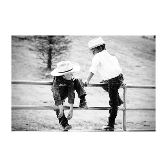 Two children in cowboy hats sitting on a metal fence outdoors, with a focus on their interaction in a monochromatic setting.