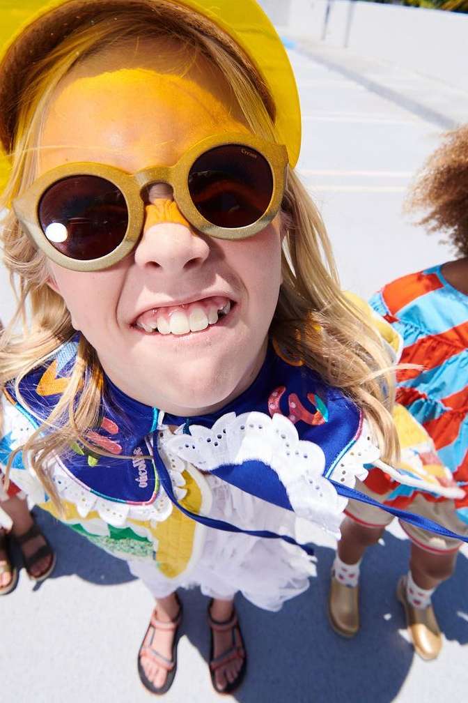 Close-up, high-angle shot of a grinning child wearing sunglasses and a yellow hat, with colorful clothing and several other children.
