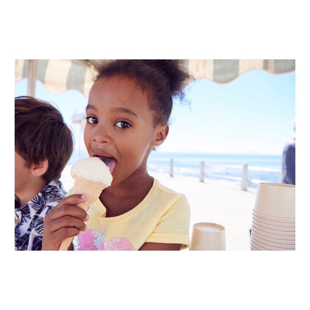 A young African-American girl at the beach, eating ice cream, looking directly at the camera with a happy expression, in daylight.