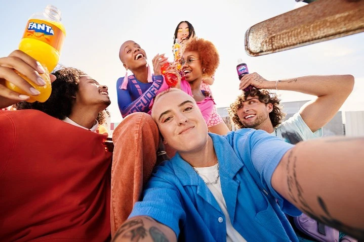 Group of diverse young adults outdoors, laughing and holding Fanta bottles, captured in a selfie-style shot under bright sky.