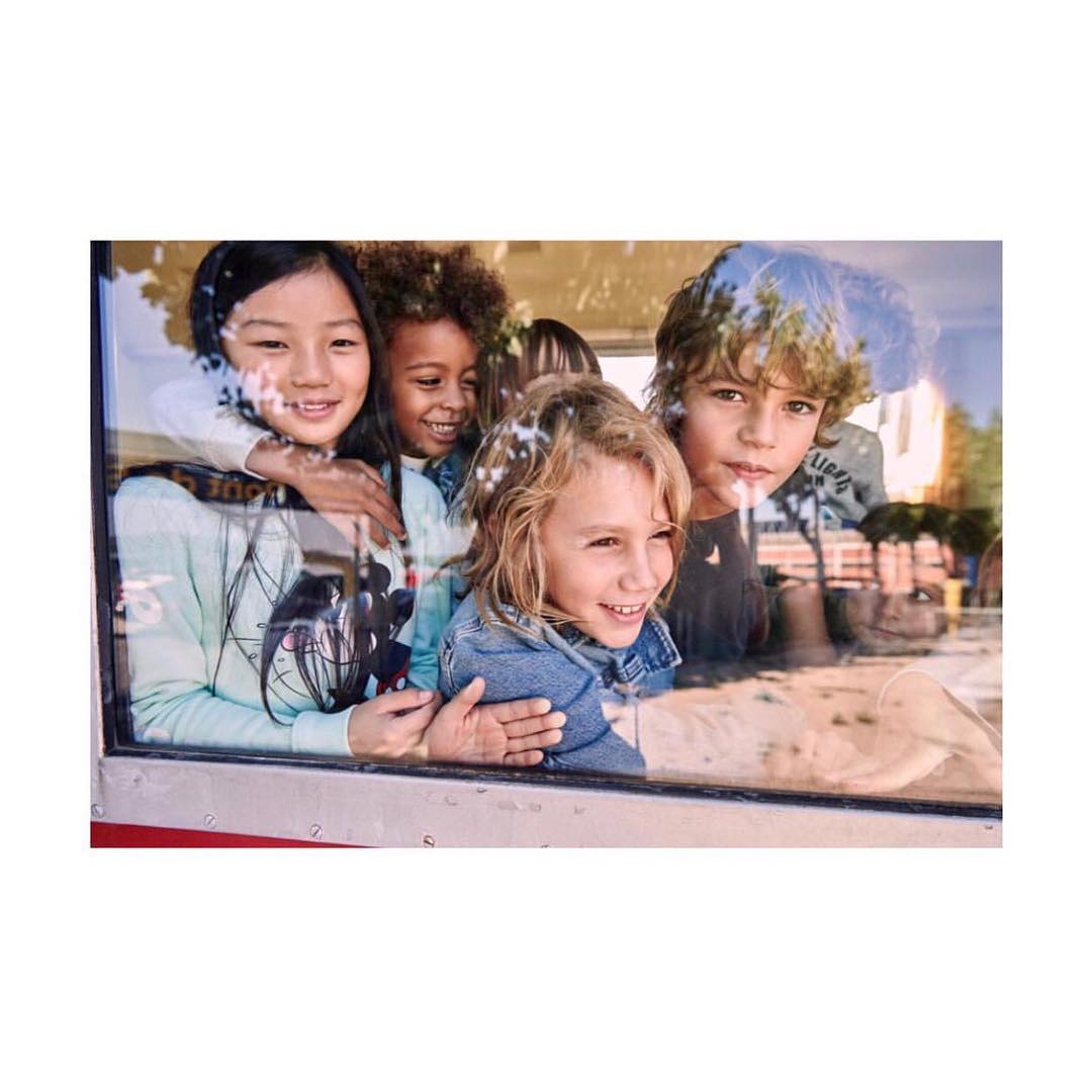A group of four multi-ethnic children are indoors smiling and looking through a window. The children are wearing casual clothing.