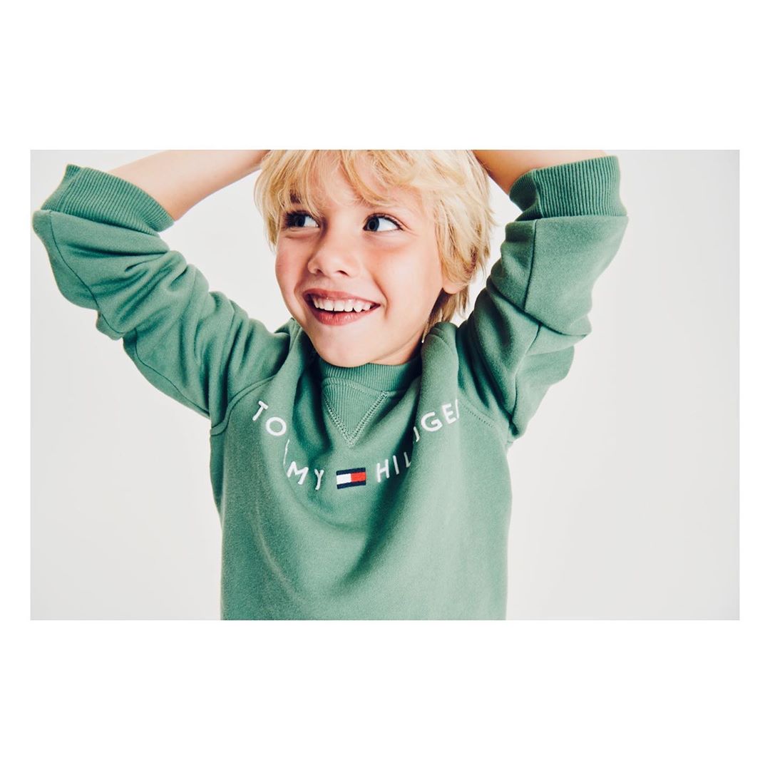 Close-up of a smiling young boy wearing a green Tommy Hilfiger sweatshirt against a plain background, showcasing children's fashion.