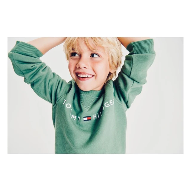 Close-up of a smiling young boy wearing a green Tommy Hilfiger sweatshirt against a plain background, showcasing children's fashion.
