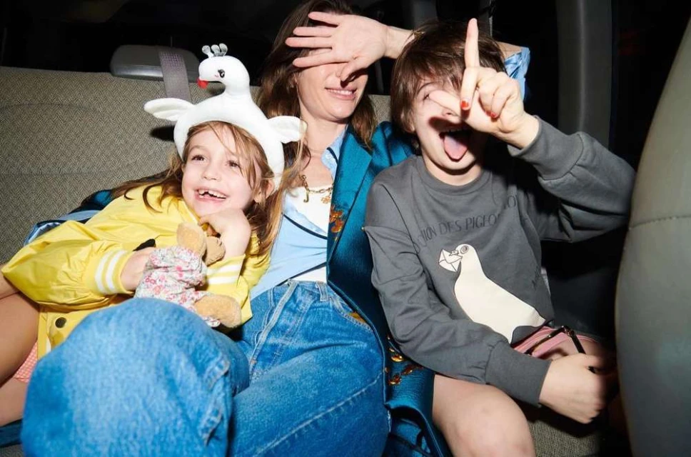 Three people - a mother with two children, a girl wearing a swan hat, and a boy making a face, sitting in a car interior setting.