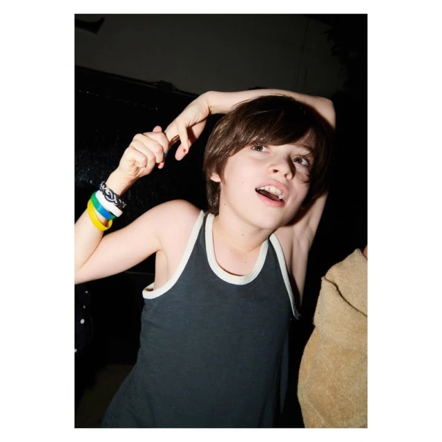 A young child with brown hair and a dark tank top raises their arms in an indoor setting, with a dark background and minimal lighting.
