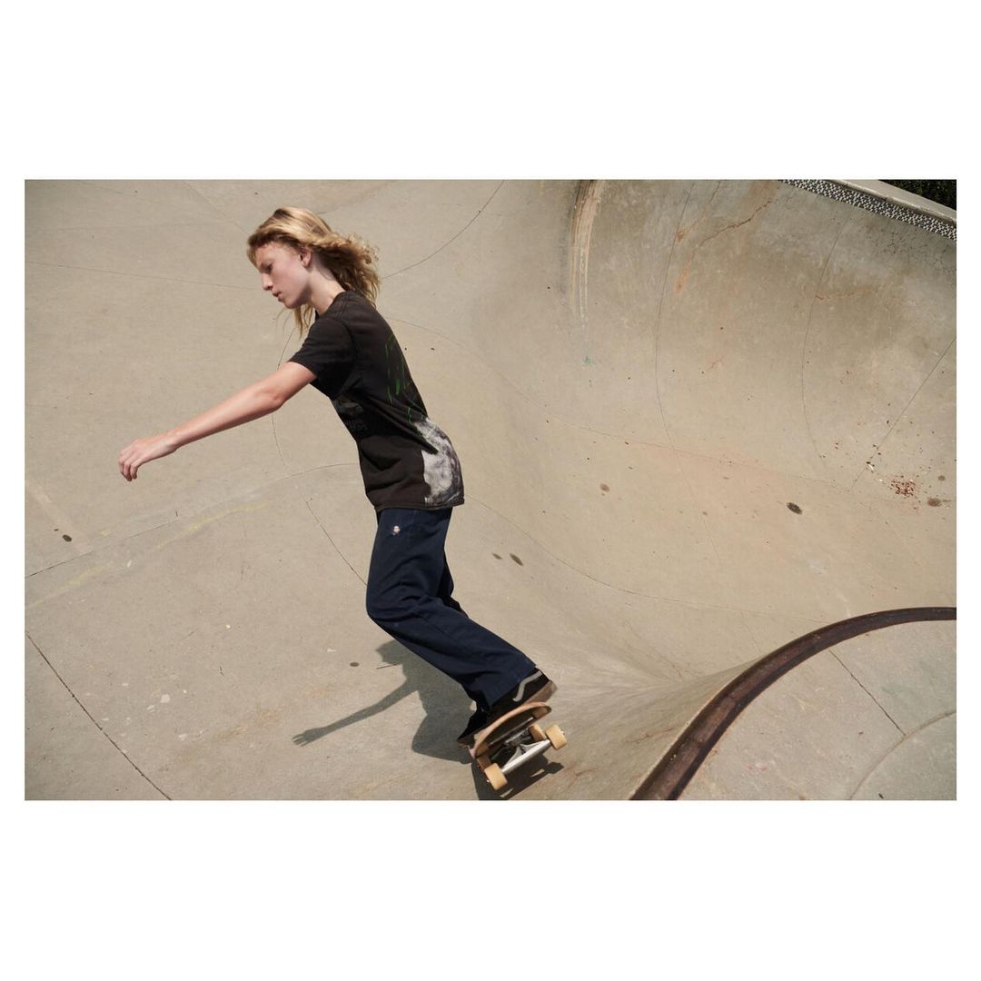 A young adult skateboarder riding a skateboard inside a concrete skatepark bowl with blonde hair, a graphic tee, and dark wash jeans.