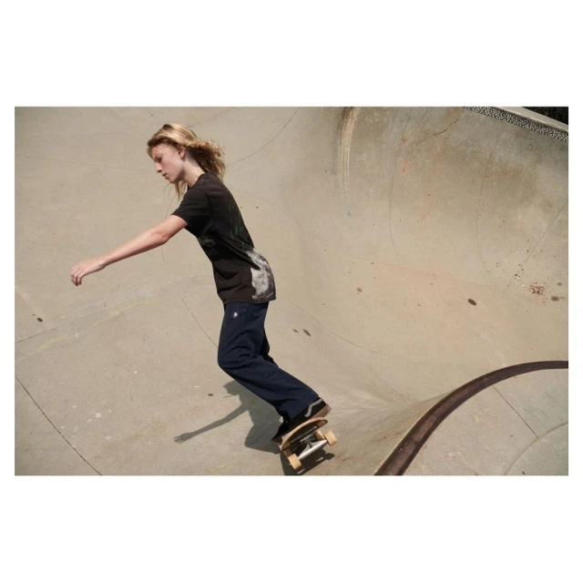 A young adult skateboarder riding a skateboard inside a concrete skatepark bowl with blonde hair, a graphic tee, and dark wash jeans.