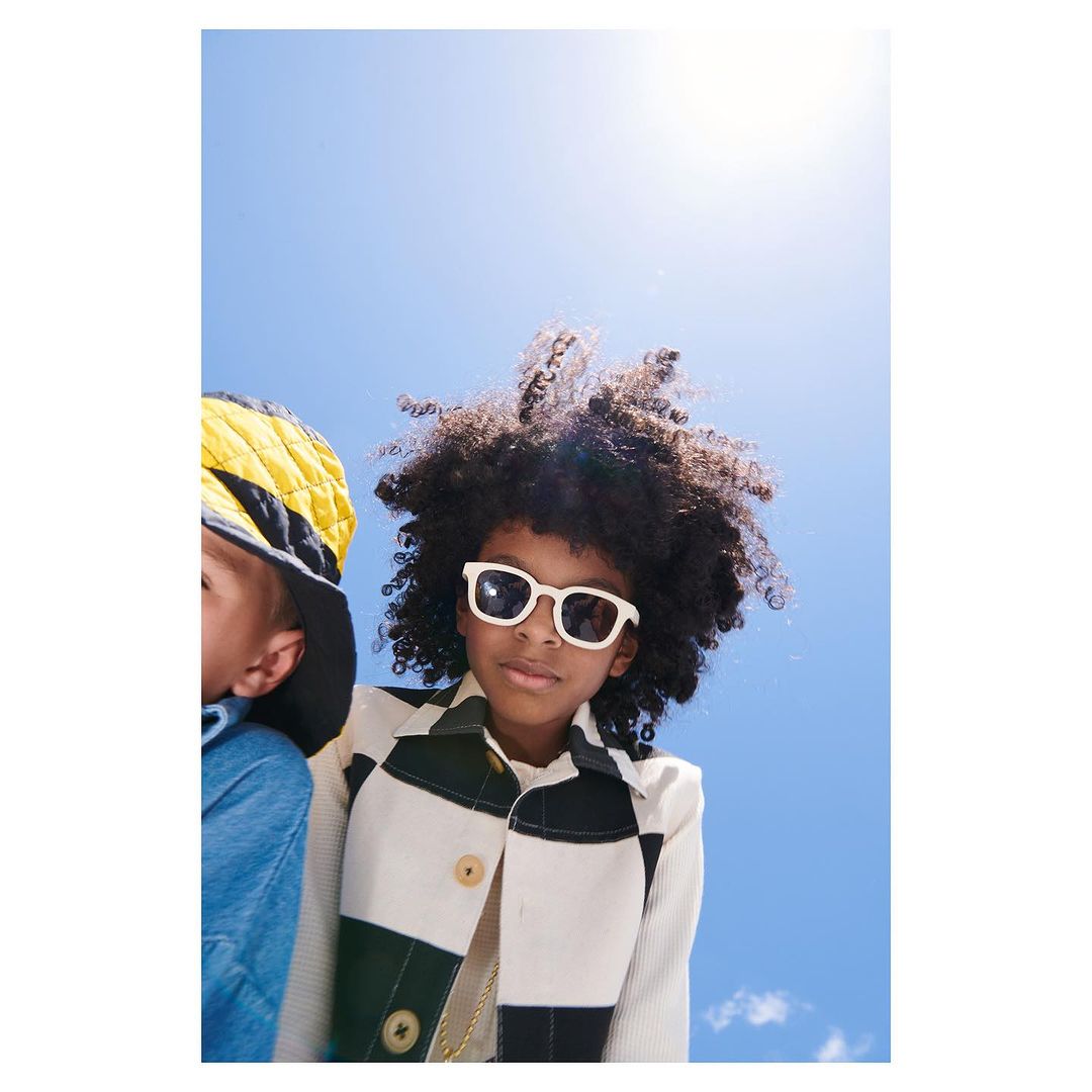 Young child poses outdoors in white sunglasses and jacket; another child with a yellow hat partially visible to the side on a clear sky day.