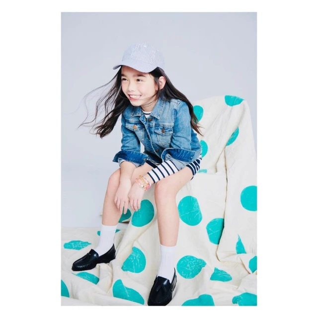 Smiling young girl in jean jacket, striped dress, and cap sitting on polka-dotted chair in a studio, cheerful and fashionable. 