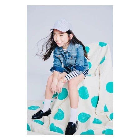 Smiling young girl in jean jacket, striped dress, and cap sitting on polka-dotted chair in a studio, cheerful and fashionable. 