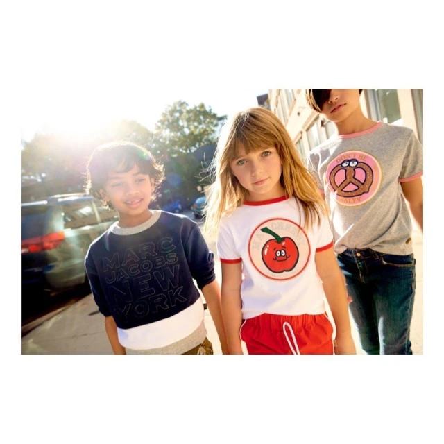 Three children stand outside on a sunny day: a boy in a Marc Jacobs sweatshirt, a girl in a red-trimmed apple tee, and a girl in a pretzel shirt.