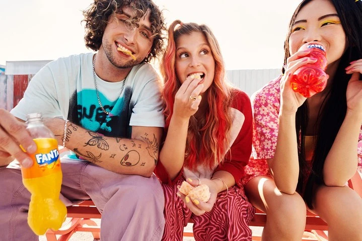 Three young adults, one man and two women, seated outdoors, sharing a refreshing orange soda and a snack on a sunny day.