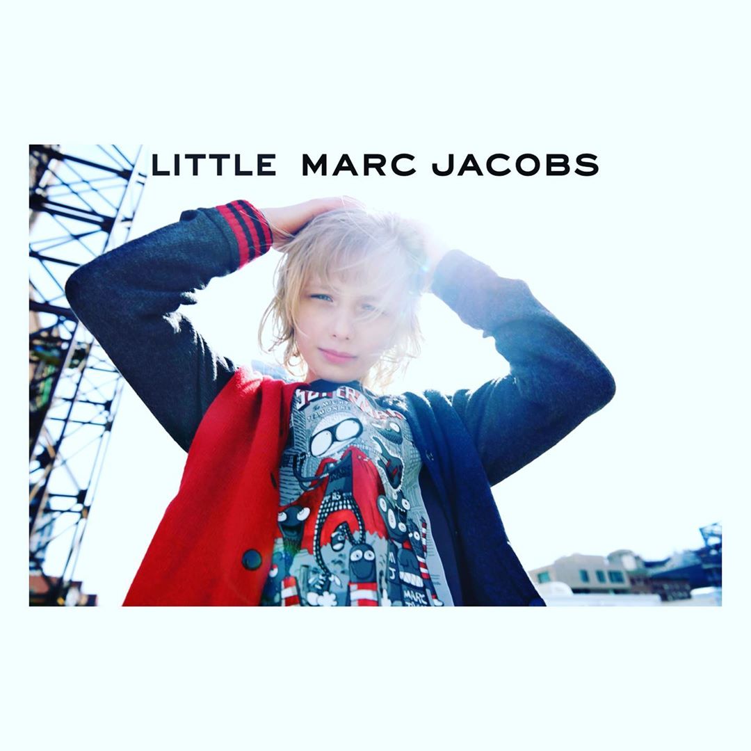 Blond child poses in Little Marc Jacobs graphic tee and jacket against a bright, low-angle urban backdrop with modern buildings.