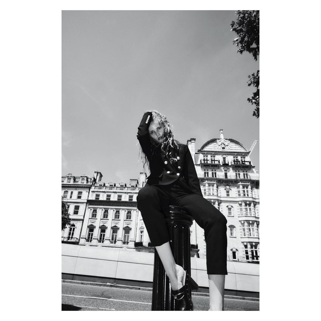 Black and white fashion portrait of a female model in a blazer and pants sitting on a bollard with city buildings in the background.