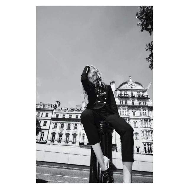 Black and white fashion portrait of a female model in a blazer and pants sitting on a bollard with city buildings in the background.