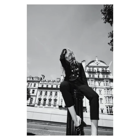 Black and white fashion portrait of a female model in a blazer and pants sitting on a bollard with city buildings in the background.