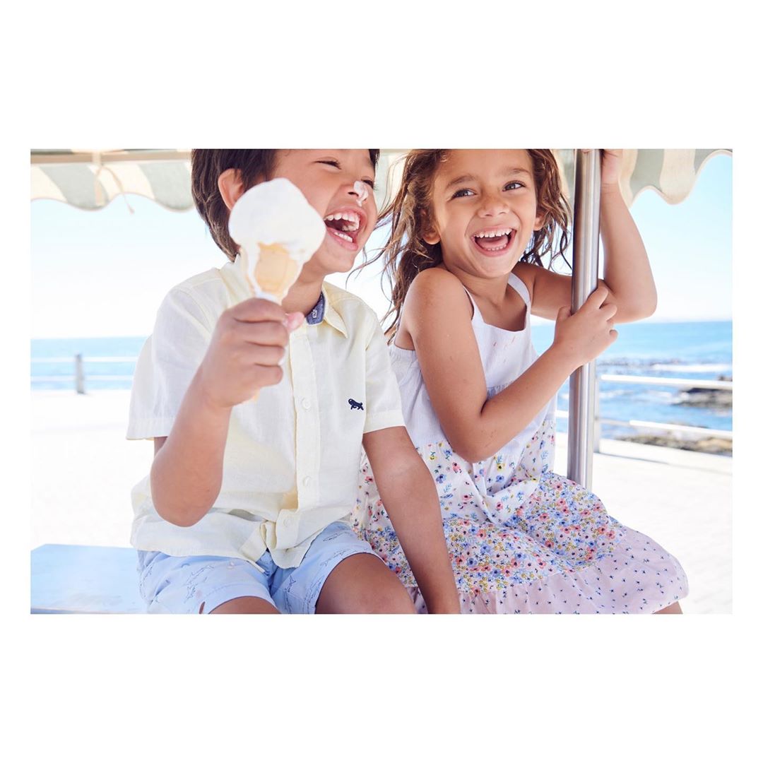 Two smiling children enjoy ice cream outdoors. A boy holds the cone, with a girl sitting nearby, with the ocean in the background.