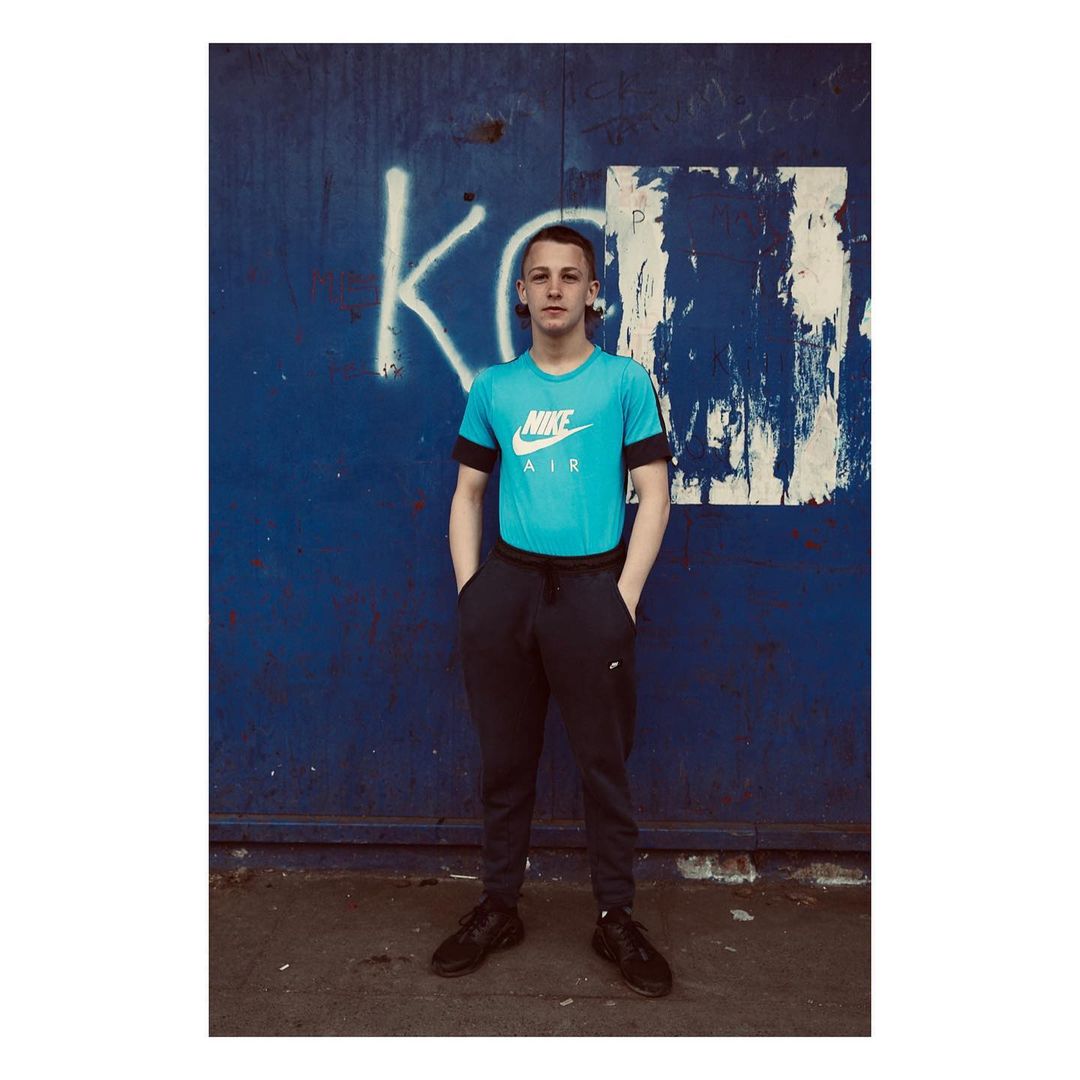 Full-length portrait of a young adult male in sportswear standing in front of a blue wall with graffiti, hands casually in pockets.