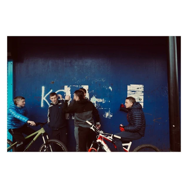 Five young men gather with their bikes in front of a blue wall with graffiti, creating a casual outdoor scene in daylight.