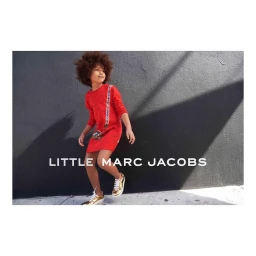 Young child with curly hair wearing a red dress and gold sneakers standing in front of a dark concrete wall