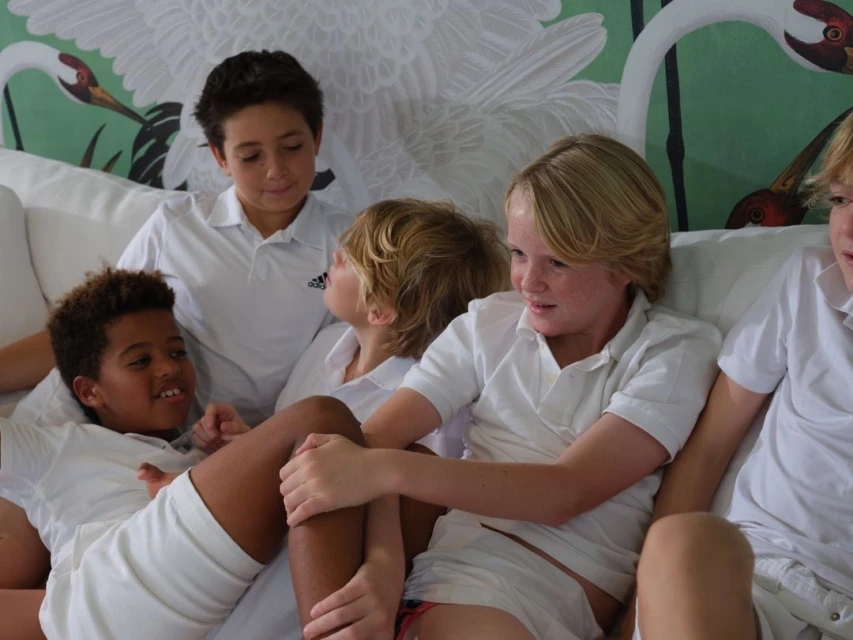 Five young boys wearing white shirts are gathered on a white couch in a well-lit interior, with a heron-patterned wall in the background.