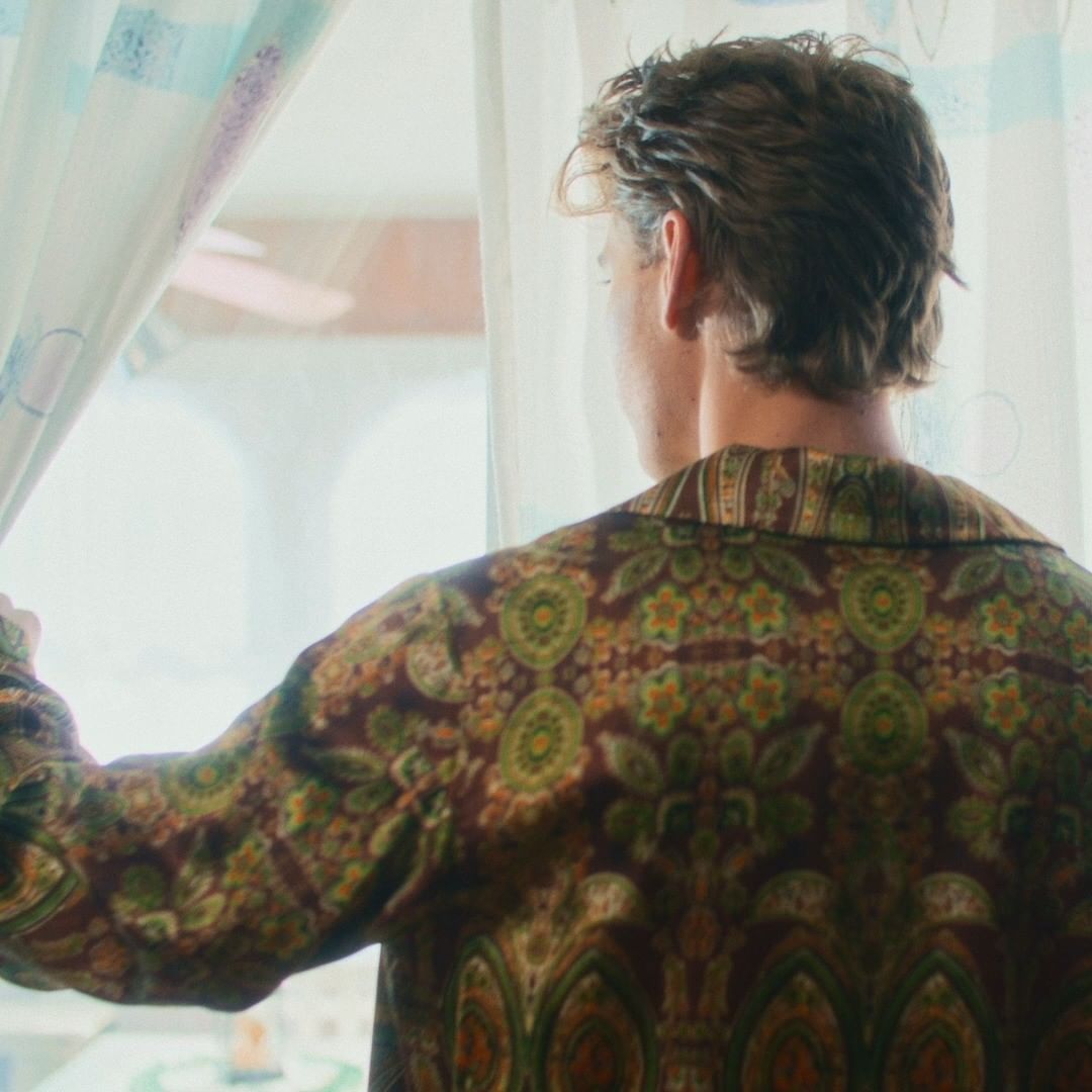Back view of a person in a patterned robe looking out a window with white curtains into a bright, out-of-focus outdoor scene.