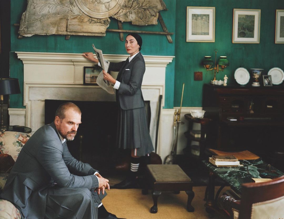 Elegant interior shot of two people wearing grey suits and kilts, one reading a newspaper, in a room with antiques and teal walls.