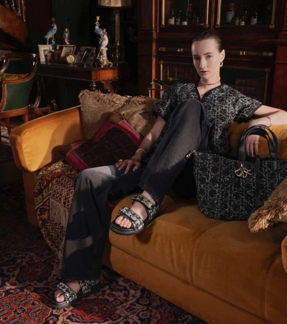 Woman poses in patterned blouse, black pants and sandals on a mustard couch next to black patterned handbag indoors with decorative background.