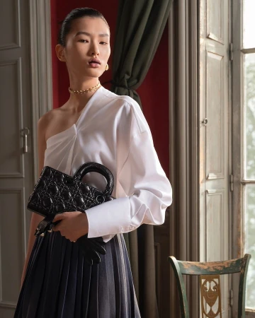 East Asian woman holding a black quilted handbag wearing an asymmetric white blouse and dark pleated skirt indoors.