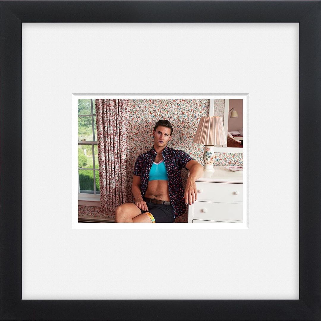 A man in an open shirt and shorts sits in a bedroom with floral wallpaper, beside a window, next to a white dresser and lamp.