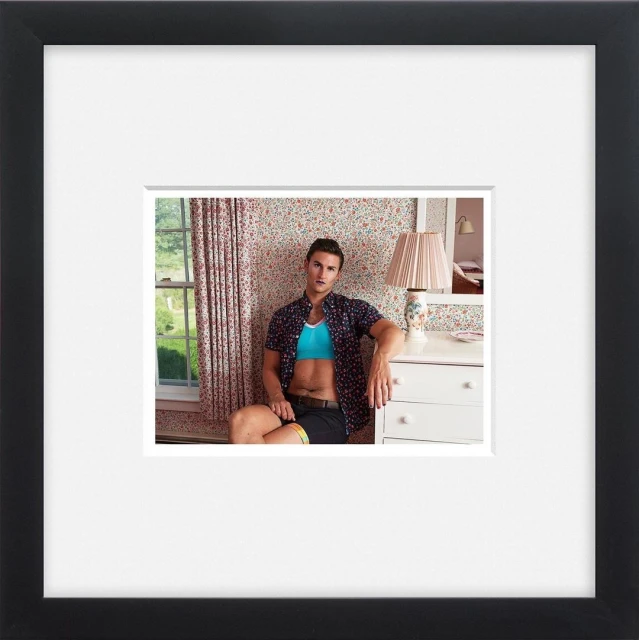 A man in an open shirt and shorts sits in a bedroom with floral wallpaper, beside a window, next to a white dresser and lamp.