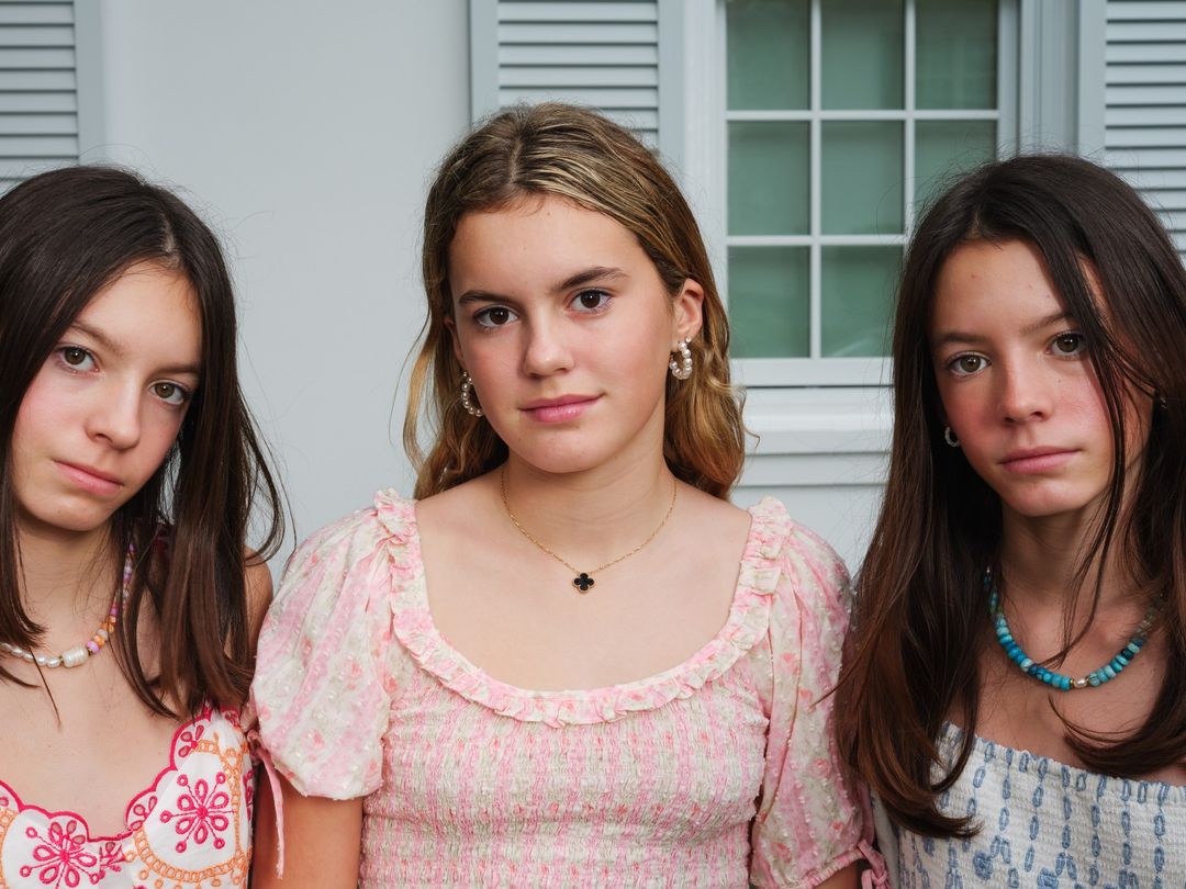Three young women pose in front of a white paneled wall. They wear summery tops and jewelry in a close-up, eye-level shot.