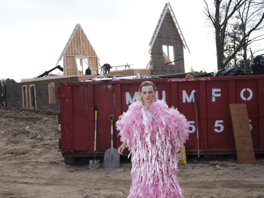 Woman wearing a pink feather boa dress stands in front of a red dumpster at a construction site with a partially built house.