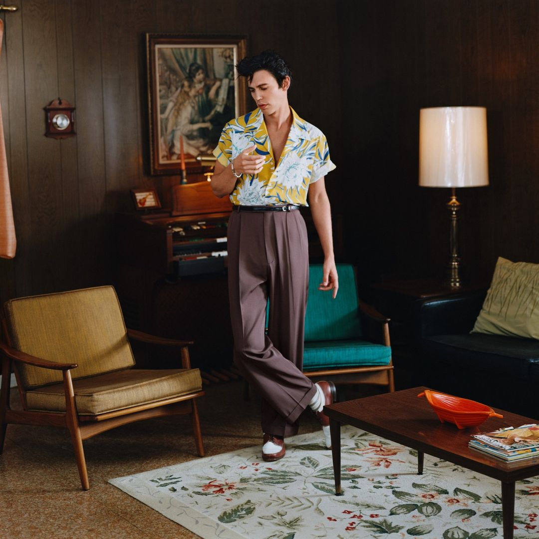 Adult male in a patterned shirt, brown slacks, and white shoes, poses in a retro-style room with a carpet, chairs, and dark wooden paneling.