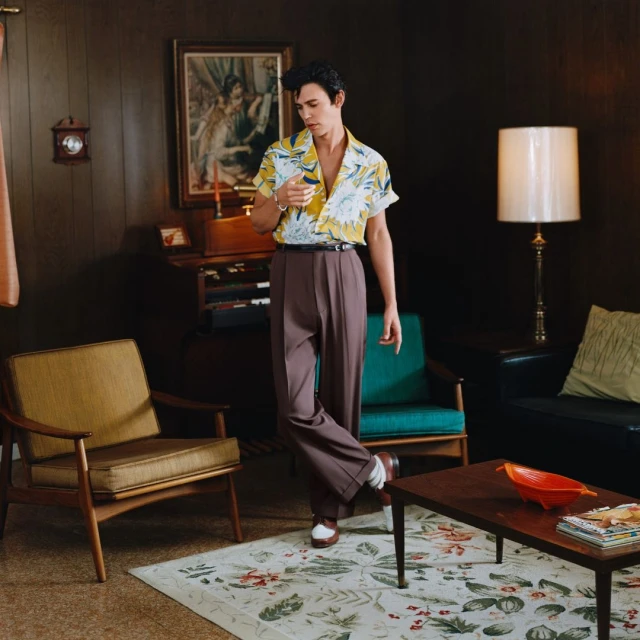 Adult male in a patterned shirt, brown slacks, and white shoes, poses in a retro-style room with a carpet, chairs, and dark wooden paneling.